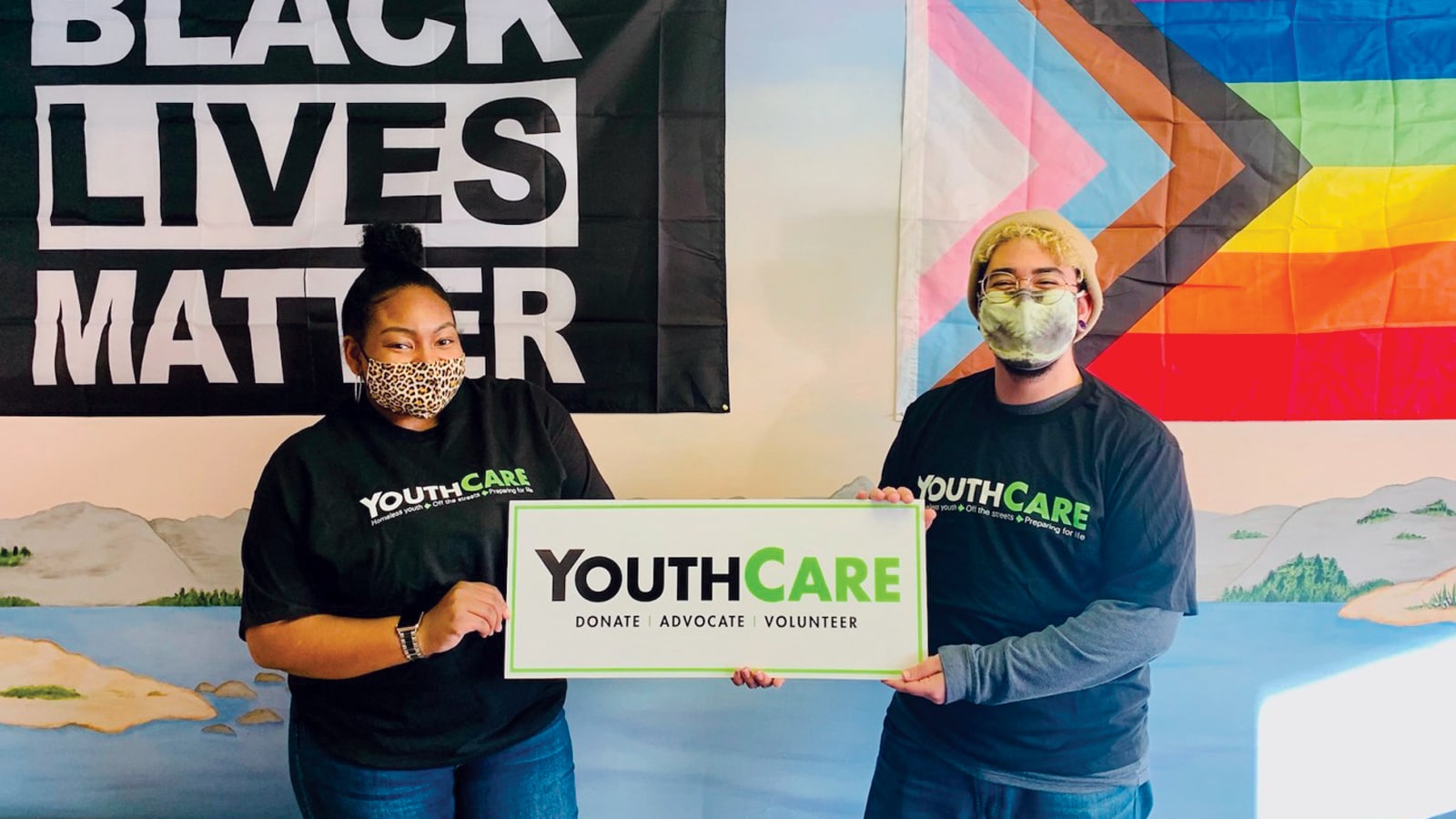 Two young people with facemasks on from YouthCare standing in front of a Black Lives Matter flag and a LGTBQ+ flag