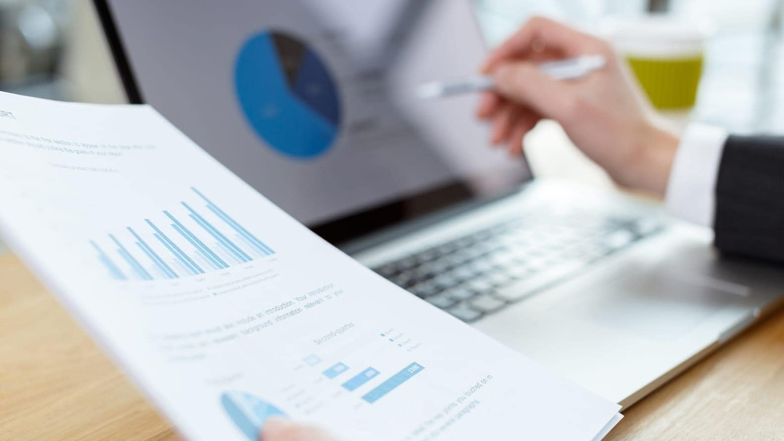 Focus image of a person reading a printed financial report and pointing to a pie graph on a laptop screen