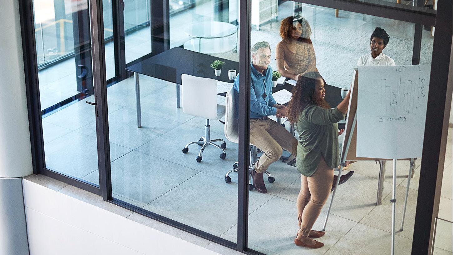 Employees in a glass conference room sitting and standing around a large notepad on an easel