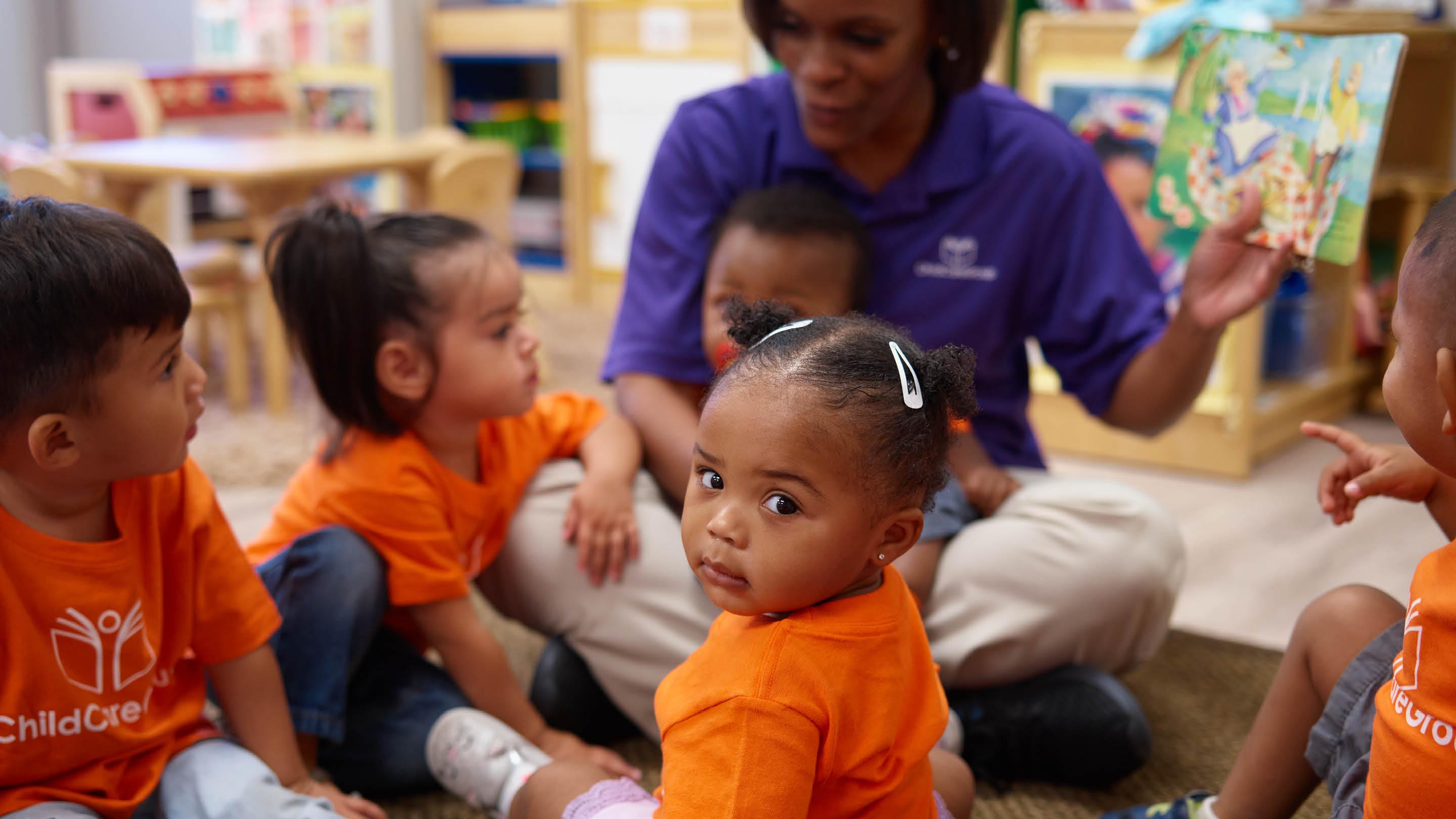 (slide 2 of 7) Young children sitting in a group with an adult teacher . 