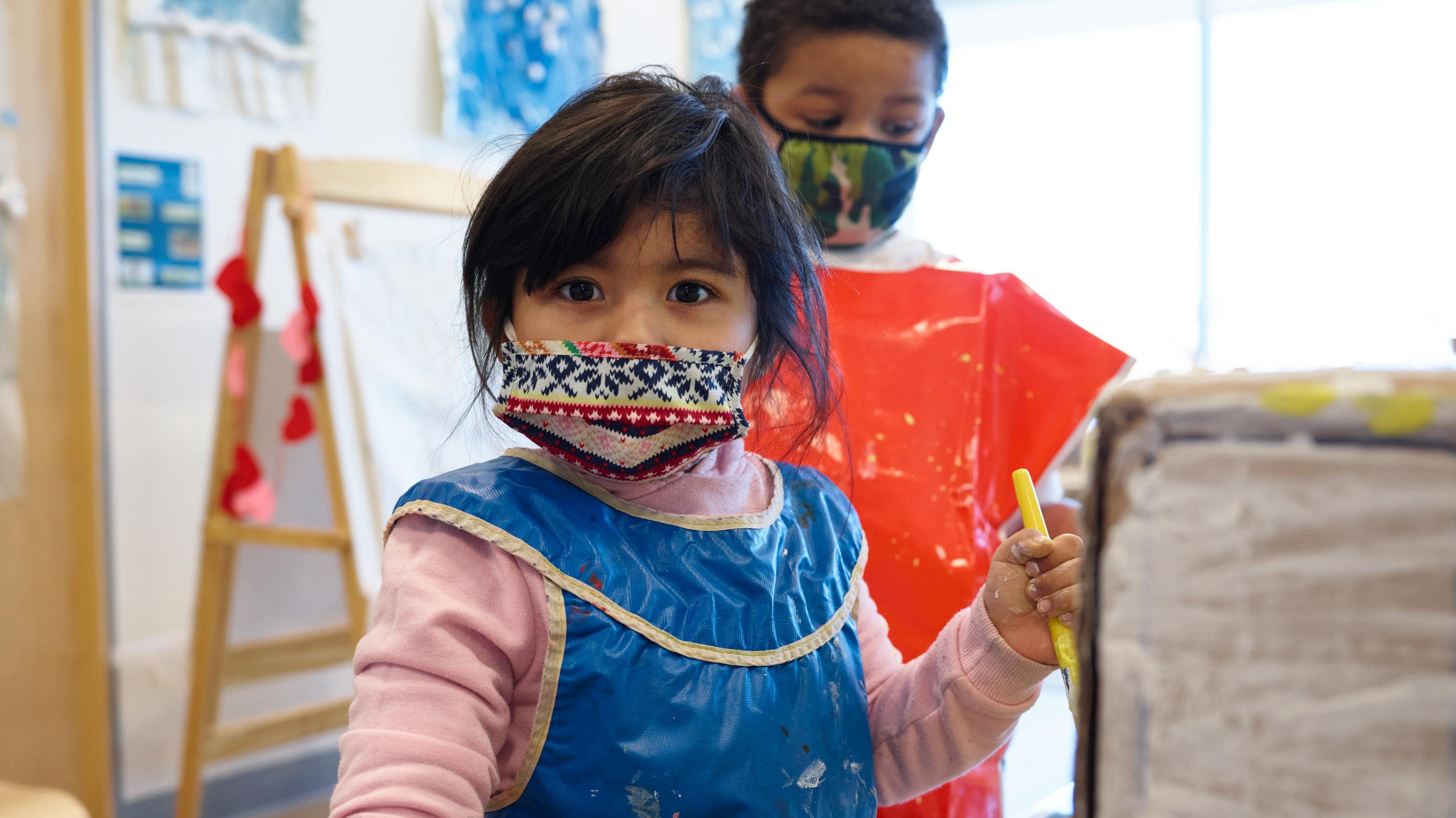 Two young children in face masks and smocks, painting and drawing in a classroom