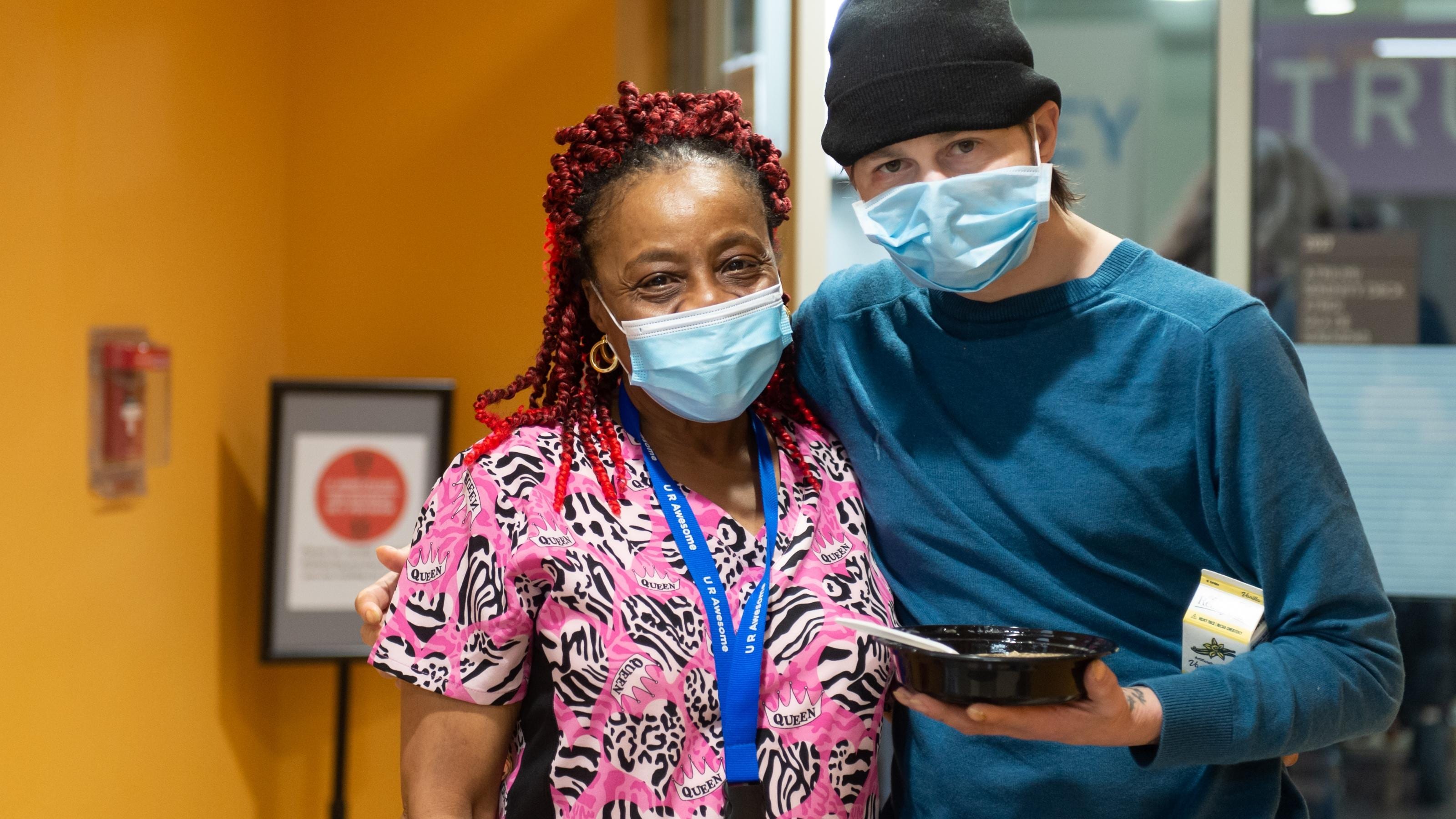 (slide 1 of 6) Two individuals at Boston Healthcare for the Homeless embracing and smiling to camera - wearing face masks. 