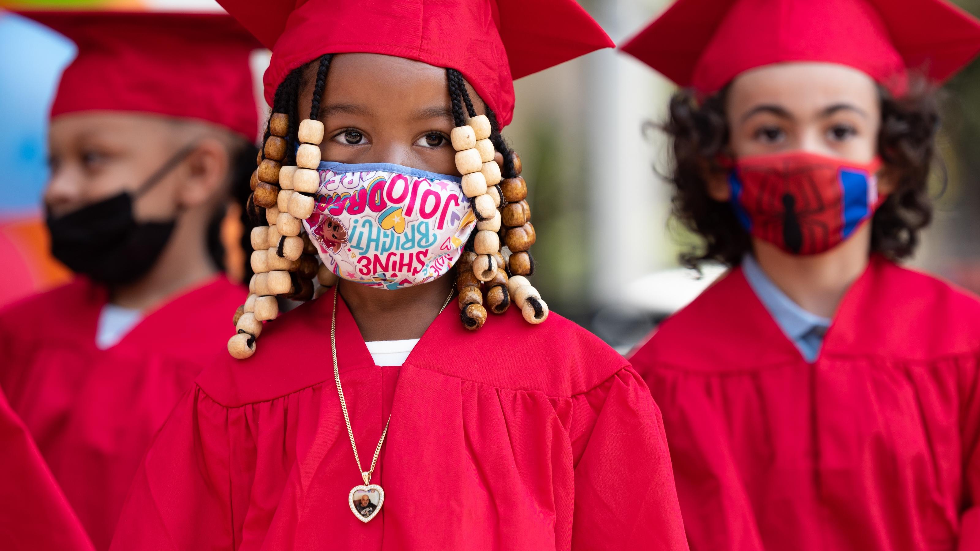 (slide 2 of 4) Young kids wearing graduation robes and face masks, lines up for graduation. 