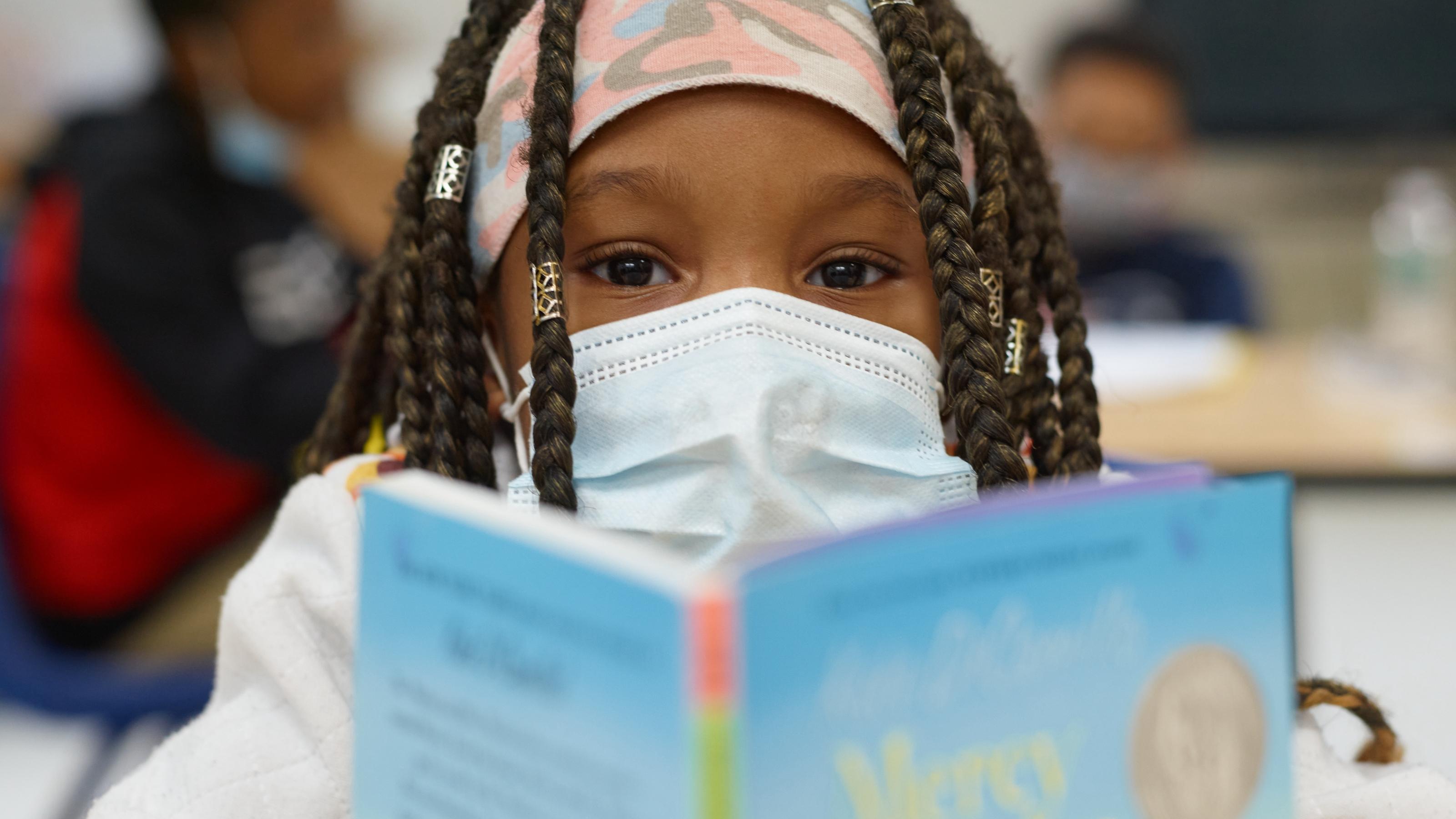 (slide 4 of 4) Young girl in a face mask reading a book in a classroom. 