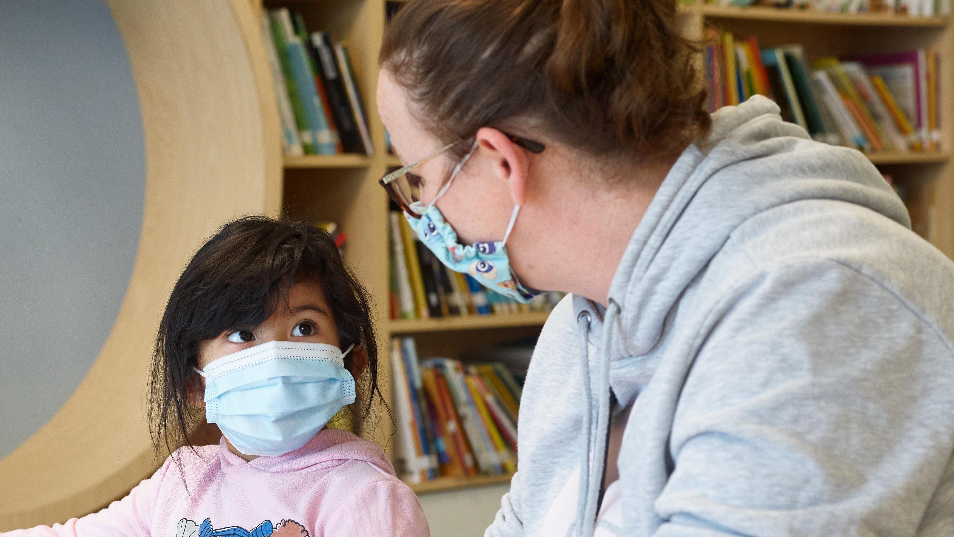 (slide 3 of 6) A masked adult, chatting with a young masked child in a classroom setting. 