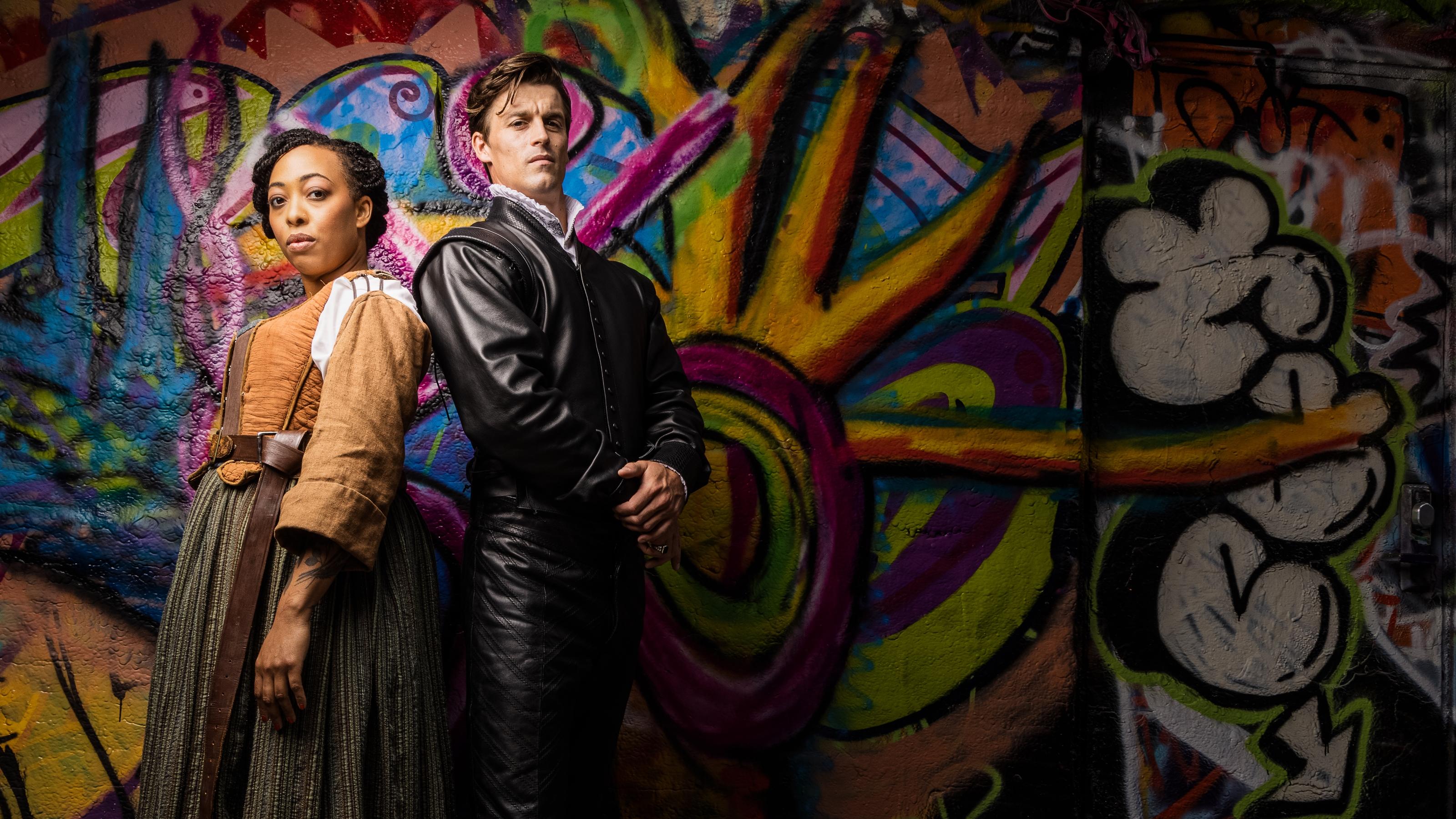 (slide 4 of 4) Woman and Man standing back to back in front of a colorful mural dressed in Shakespeare play costumes . 