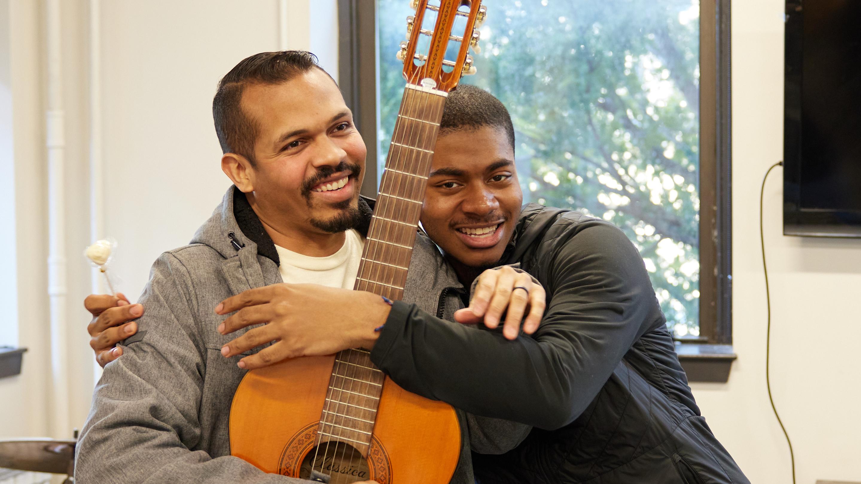 (slide 3 of 7) Two young men embracing while holding a guitar . 