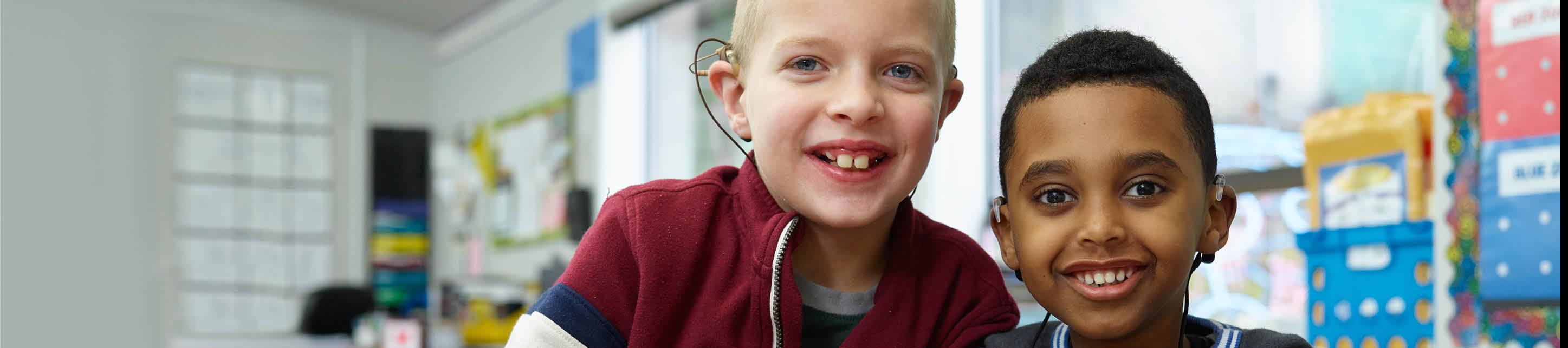 Two young children with ear implants smiling in a classroom