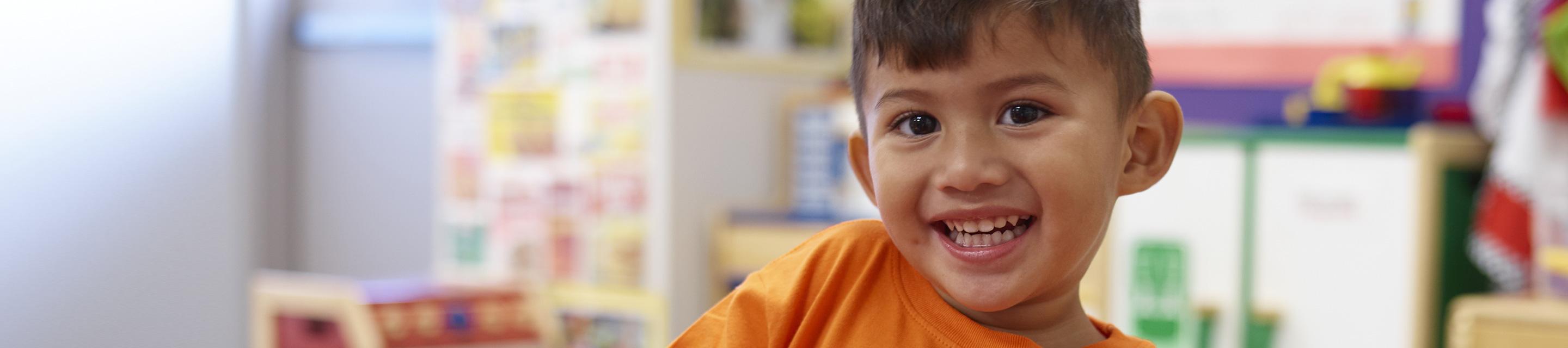 Young boy wearing an orange shirt, in a classroom, smiling to camera