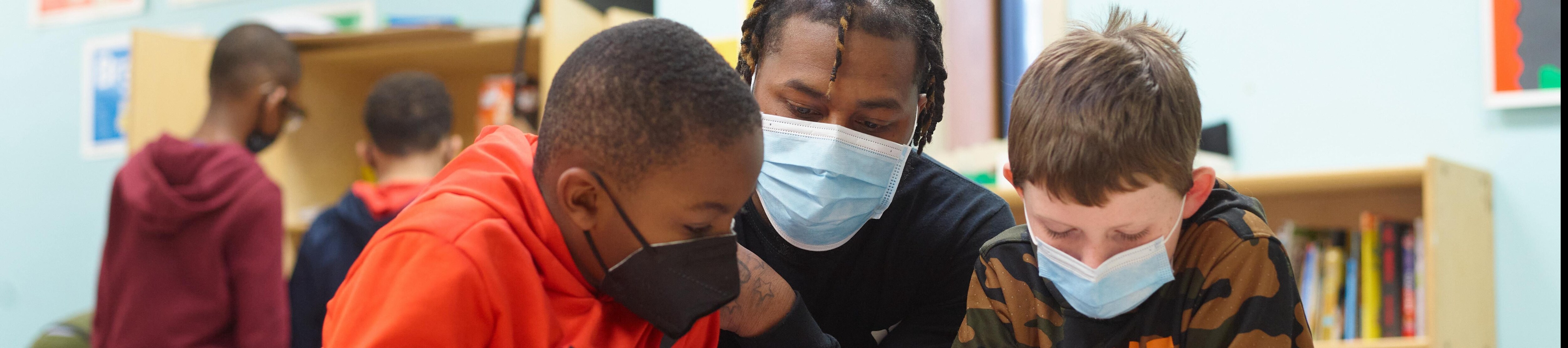 A young man and two children wearing face masks, reading a book in a classroom setting